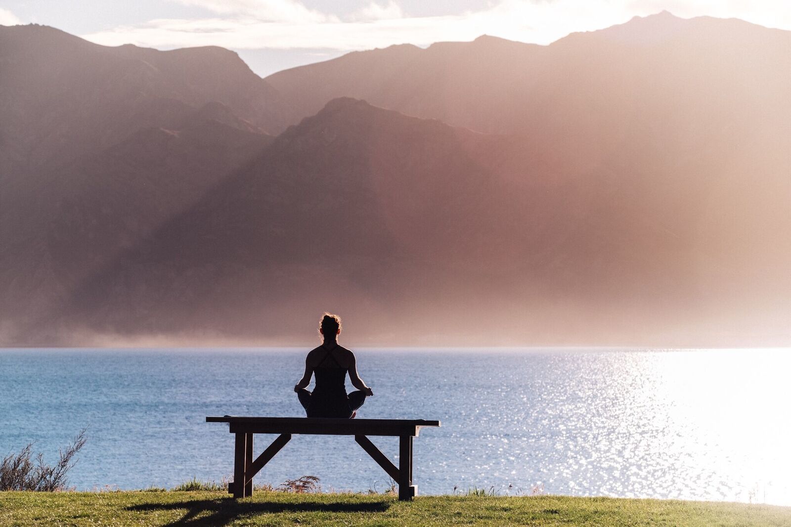 Uma pessoa meditando num banco à beira de um lago, com montanhas ao fundo.