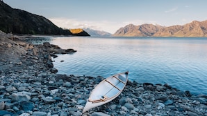 A rowboat on a rocky shore with a calm lake and mountains in the background. - The Camp - Lake Hawea (Lake Hawea)