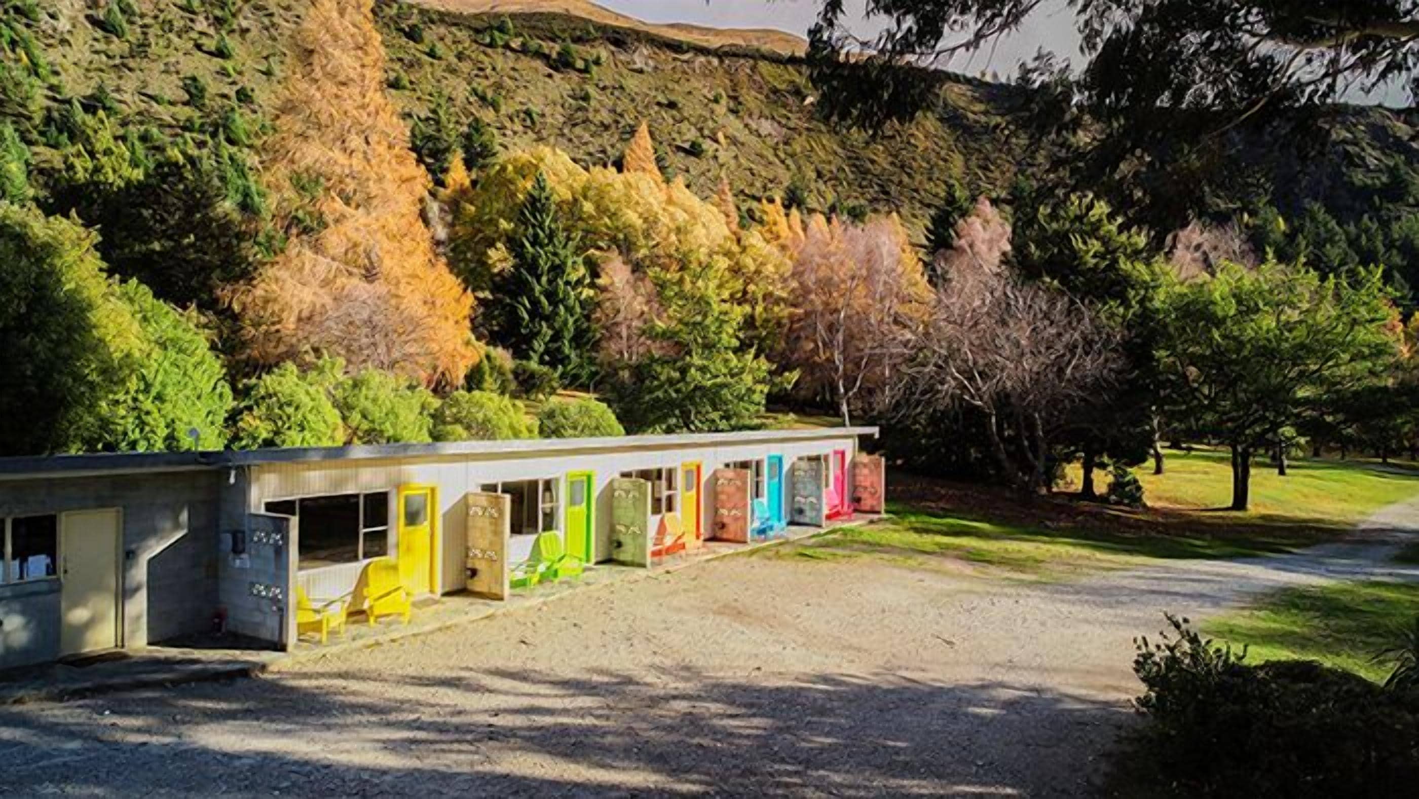 Un motel con una hilera de puertas coloridas, un camino de grava y un paisaje montañoso boscoso al fondo.