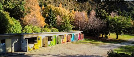 Un motel con una hilera de puertas coloridas, un camino de grava y un paisaje montañoso boscoso al fondo.