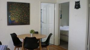 A dining area with a round table, four chairs, a peacock painting, and a door leading to a bedroom. - Willow Dene (Bright)