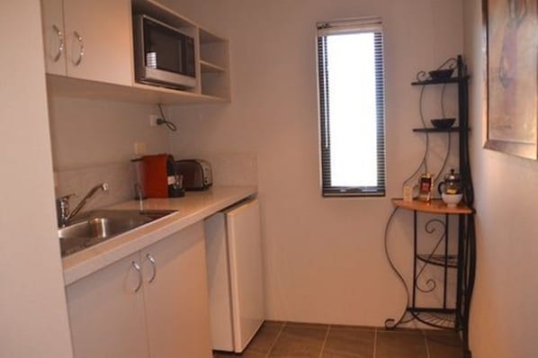 A compact kitchen with a microwave, sink, and a small refrigerator. There is a window with blinds and a shelf with kitchen items.