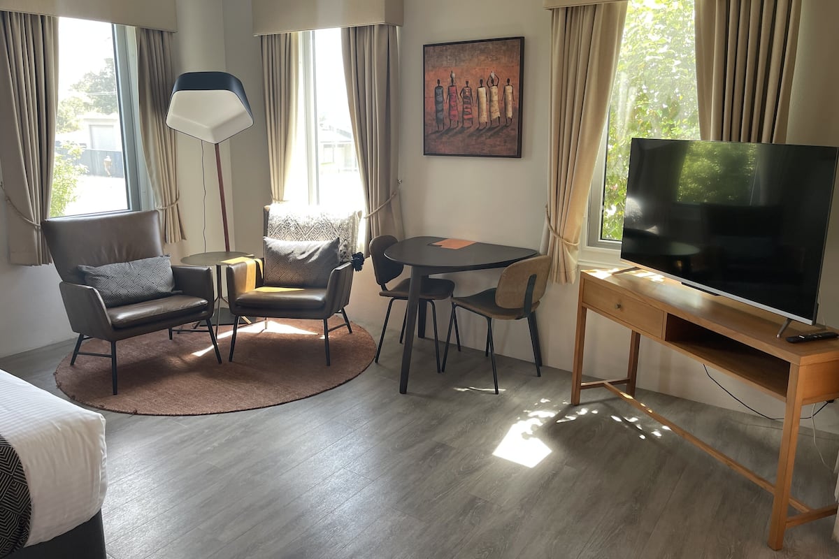 Ein modernes Hotelzimmer mit einem großen Flachbildfernseher, einem runden Esstisch, zwei Sesseln, einer Stehlampe und einem Gemälde an der Wand.