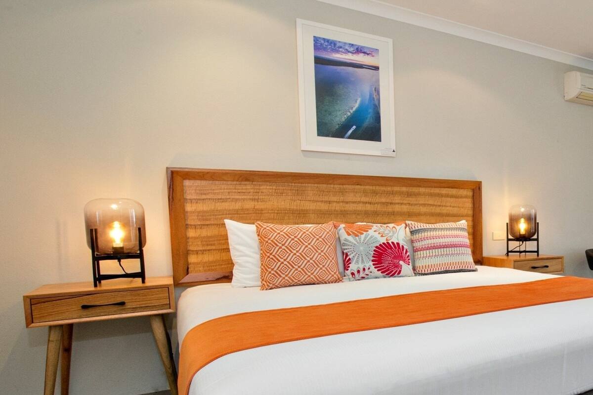 Ein ordentlich bezogenes Bett mit einer orangefarbenen Tagesdecke, einem hölzernen Kopfteil und mehreren gemusterten Kissen. Es gibt zwei Nachttische mit Lampen und ein gerahmtes Bild an der Wand.