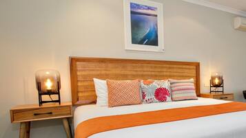 Una cama bien hecha con una colcha naranja, un cabecero de madera y varios almohadones estampados. Hay dos mesitas de noche con lámparas, y un cuadro enmarcado en la pared.