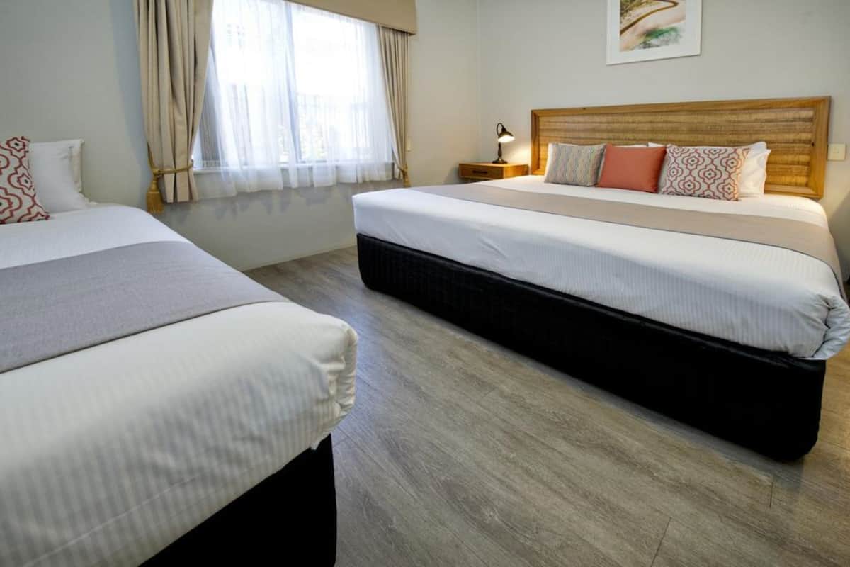 Ein Hotelzimmer mit zwei Betten, einem Holzkopfteil, einem Nachttisch mit Lampe und einem Fenster mit Vorhängen.