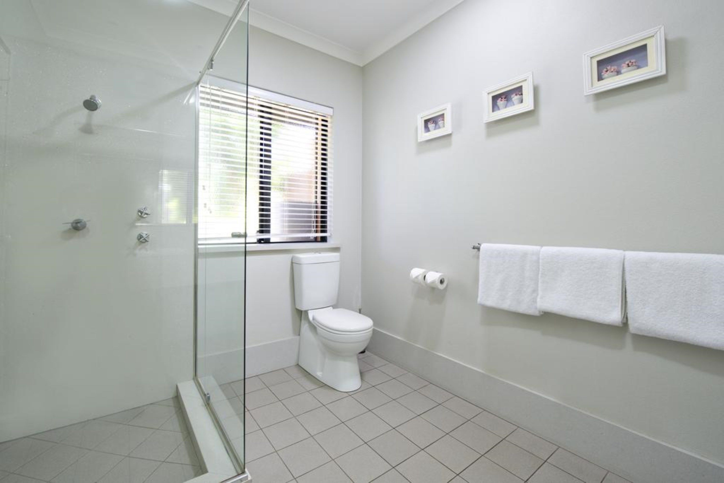 Un baño con cabina de ducha de vidrio, inodoro, ventana con persianas y toallero con toallas.