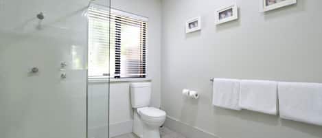 Un baño con cabina de ducha de vidrio, inodoro, ventana con persianas y toallero con toallas.