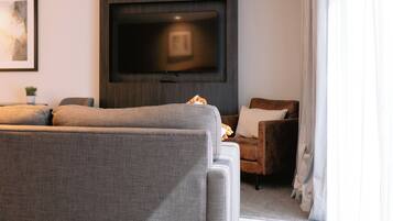 A modern hotel room with a large flat-screen TV mounted on the wall, a grey sofa, a small armchair, and a striped rug.