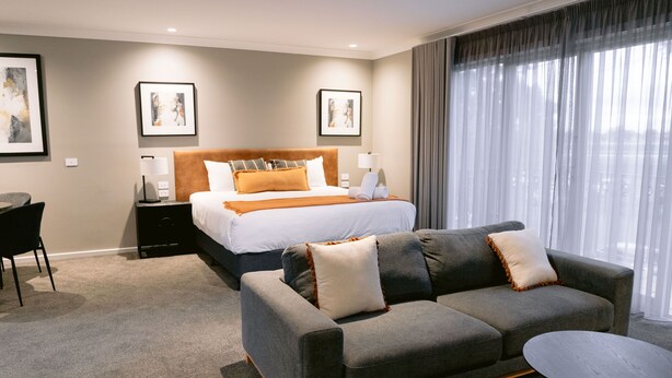 A hotel room with a bed, a grey sofa, a small table, and a desk. There are framed pictures on the wall and a window with curtains. - Rsl Club Motel (Wagga Wagga)