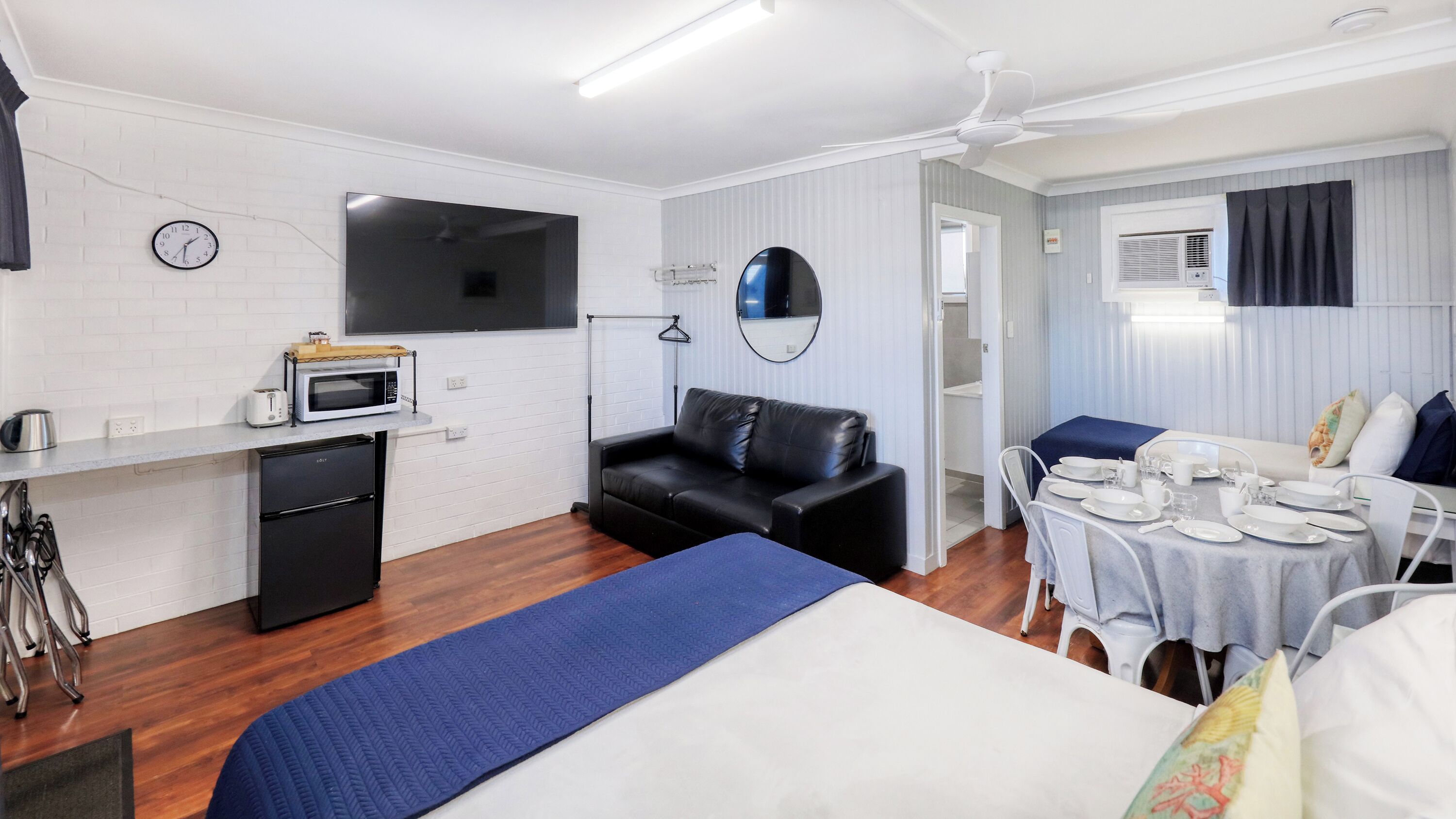 A compact hotel room with a bed, a small kitchenette, a seating area with a sofa, and a dining table set for two.
