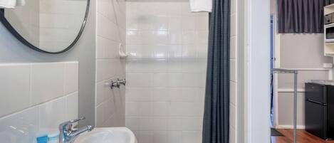 A bathroom with a white sink, a shower, and a mirror. There is a black appliance in the corner.
