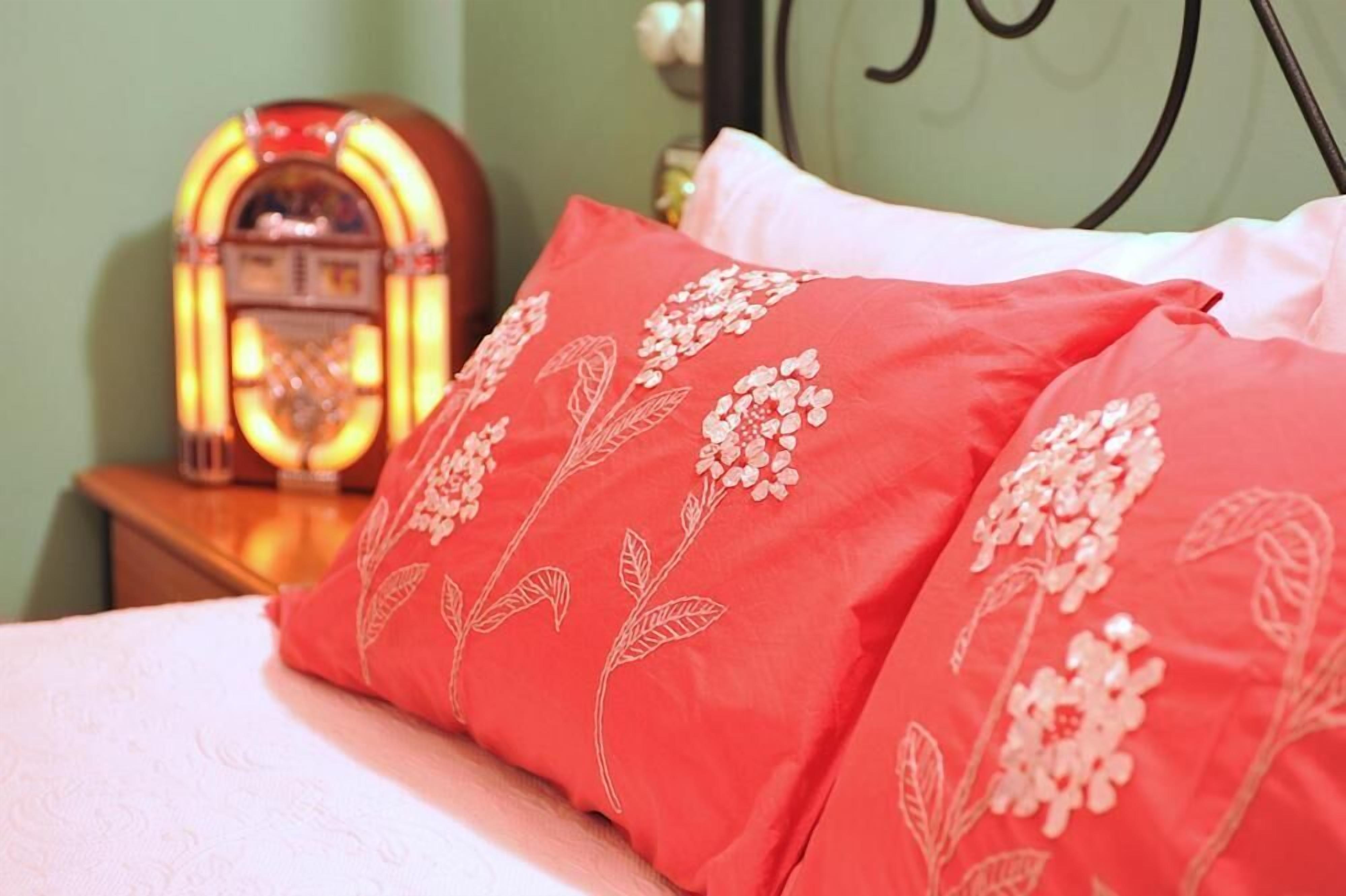 A bed with red pillows featuring floral embroidery, a vintage radio on a bedside table, and a green wall with a black metal headboard.
