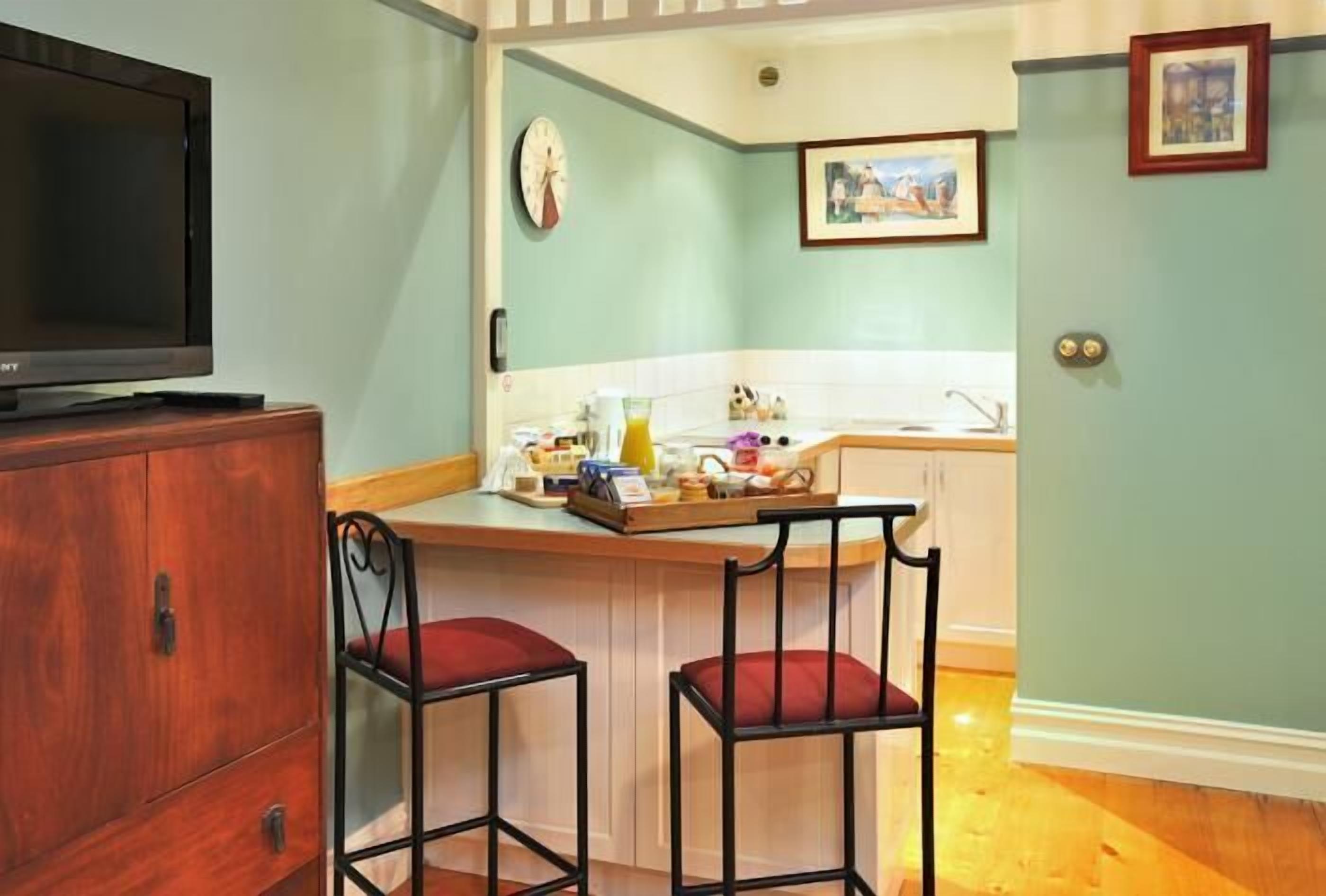 A compact kitchen with a wooden cabinet, a bar stool, a television, and a small island with a sink and various kitchen items.