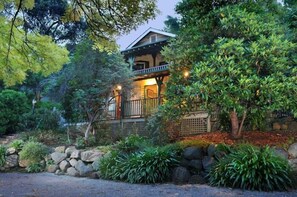 Una casa de dos pisos con balcón, rodeada de abundante vegetación y un muro de piedra.