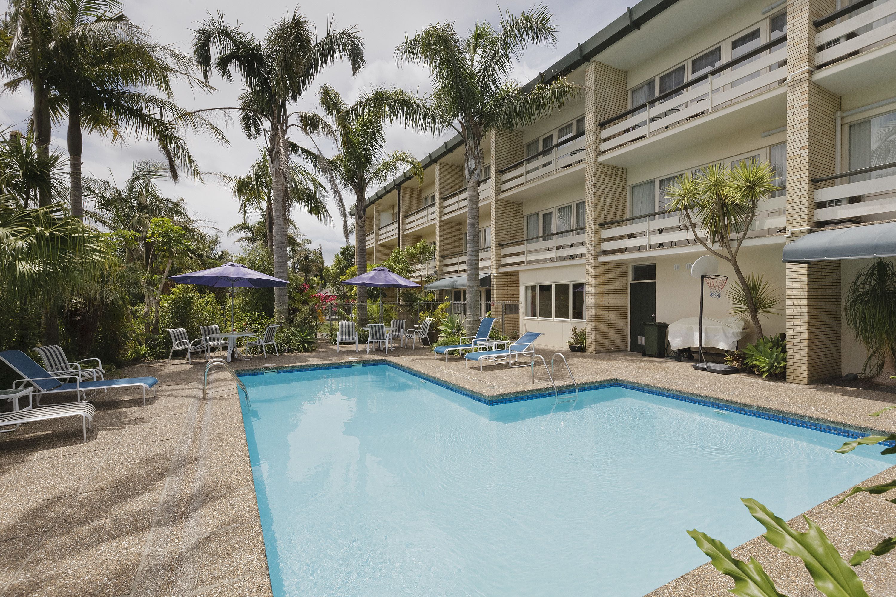 A pool area with lounge chairs and umbrellas, surrounded by palm trees and a multi-story building.