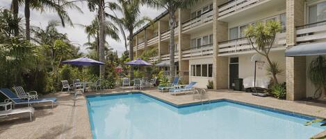 A pool area with lounge chairs and umbrellas, surrounded by palm trees and a multi-story building.