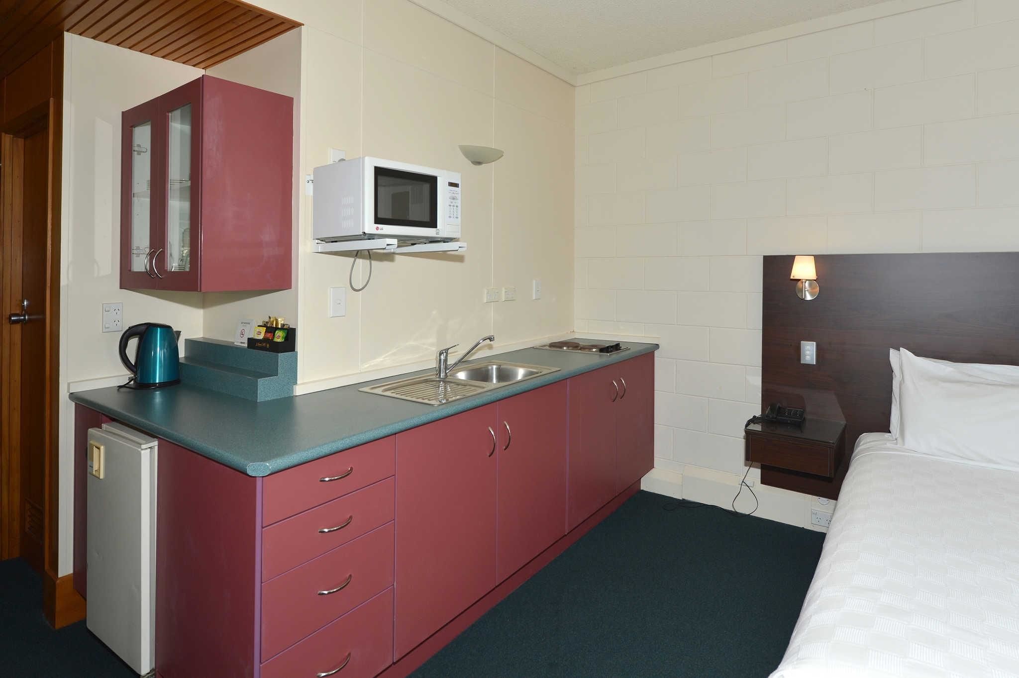 A compact hotel room with a kitchenette, a microwave, a sink, and a bedside table with a lamp.