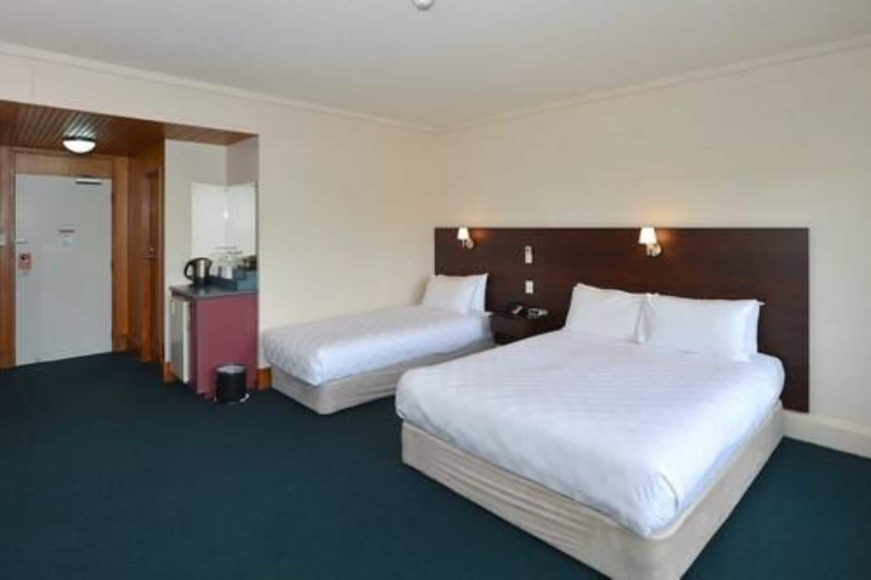 Habitación de hotel con dos camas individuales, un escritorio pequeño con una silla, un espejo y una puerta que da a otra habitación.