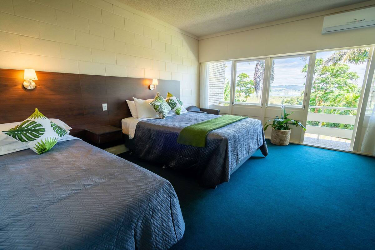 A hotel room with two beds, a large window with a view, and a potted plant.
