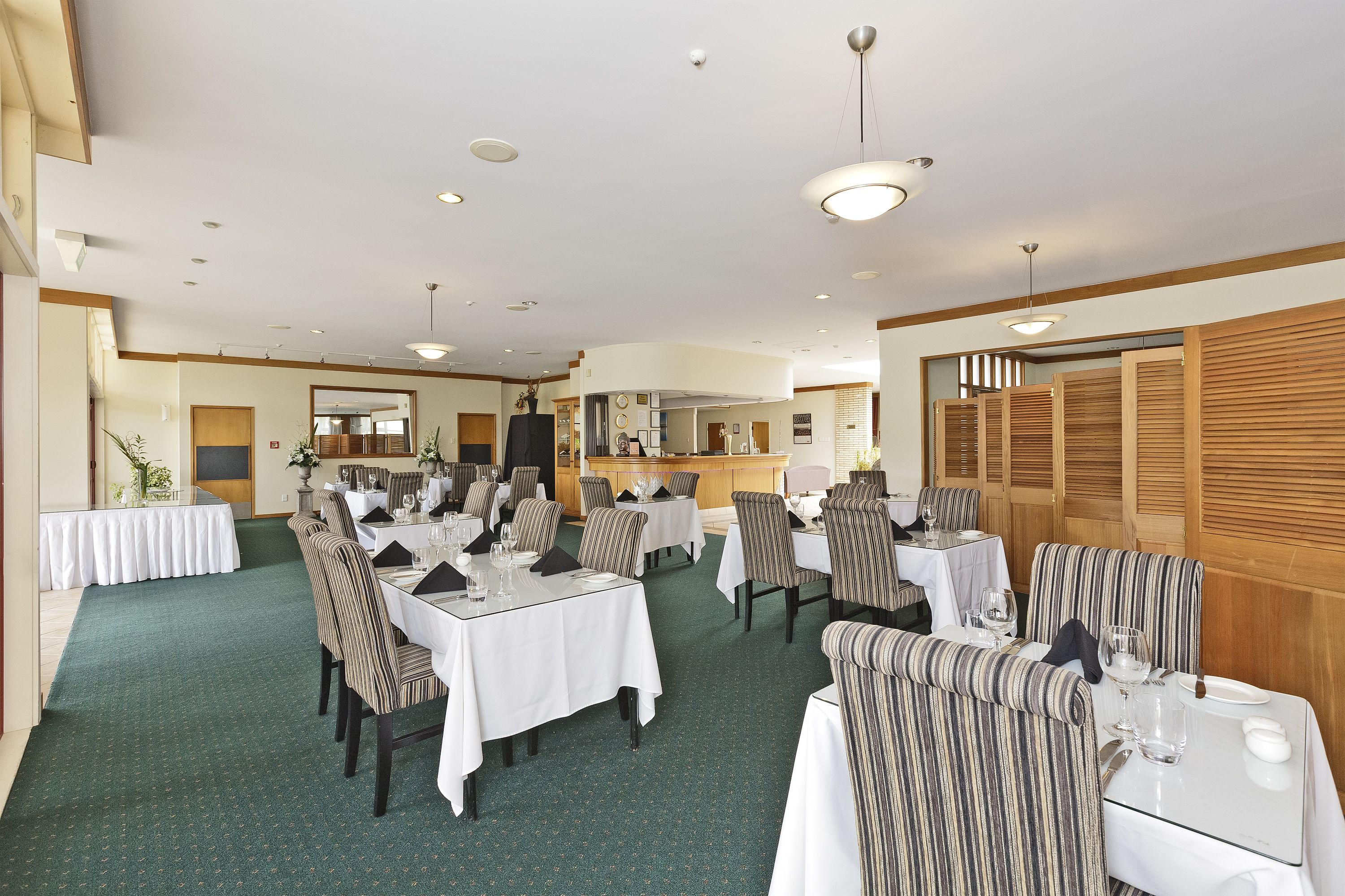 A spacious dining area with multiple tables and chairs, a kitchen in the background, and wooden paneling on the walls.