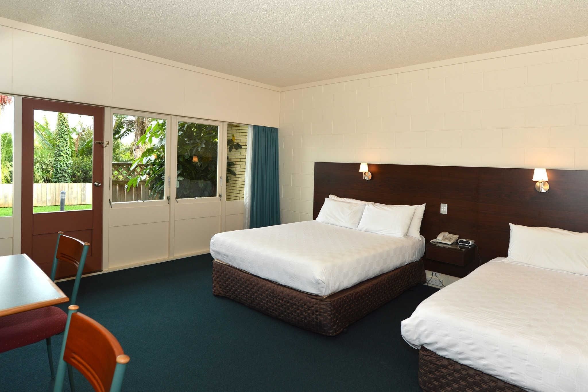 A hotel room with two beds, a desk, and a chair. There is a sliding door leading to a balcony with a view of greenery.