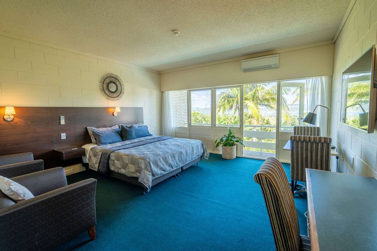 A hotel room with a bed, two bedside tables, a desk, a chair, a potted plant, and large windows with a view of palm trees.