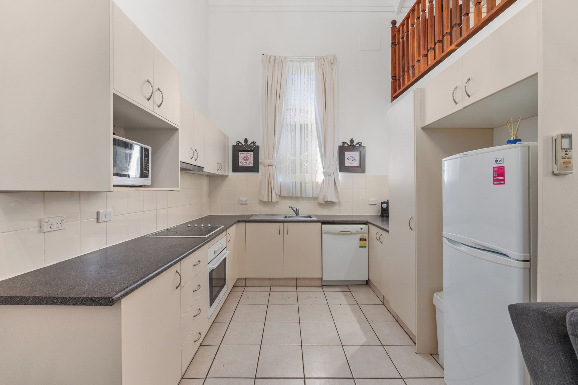 A compact kitchen with a microwave, oven, and refrigerator, a window with curtains, and a wooden staircase.