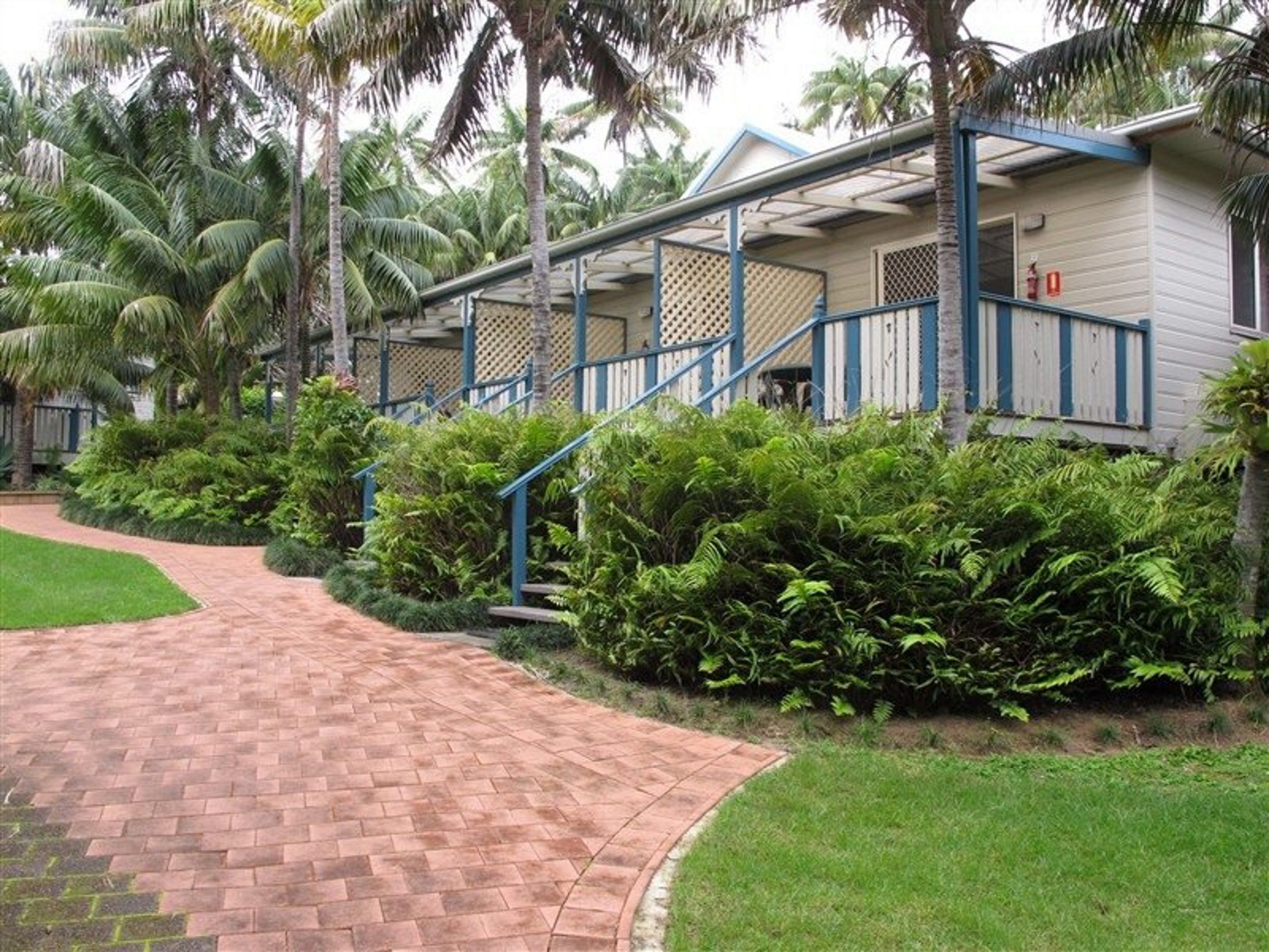 Ein Bungalow mit Ziegelweg, der zu einer Holterrasse führt, umgeben von üppiger Vegetation und Palmen.