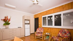 A compact living space with a kitchenette, two wicker chairs, a dining table, and a window with blinds.