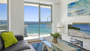 A modern living room with a large window overlooking a beach, a flat-screen TV, and a glass coffee table. - Foreshore Apartments (Mermaid Beach)