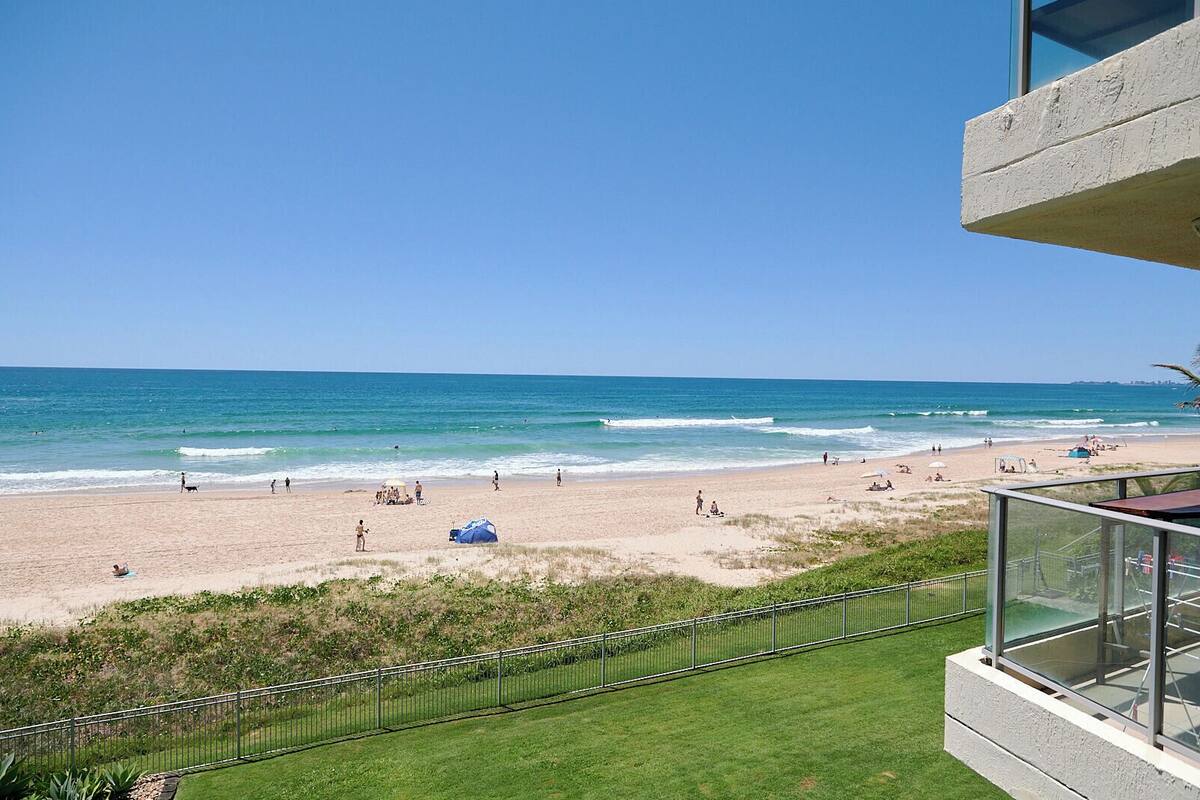A beach view from a balcony with a clear sky, a sandy beach, and a few people and umbrellas.