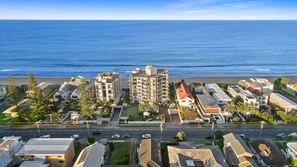 Un hôtel situé sur la côte, composé de plusieurs bâtiments, donnant sur la plage et bordé d’une route où circulent des voitures.