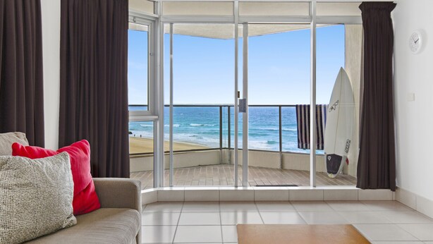 A living room with a view of the beach, a surfboard, and a clock on the wall. - Foreshore Apartments (Mermaid Beach)