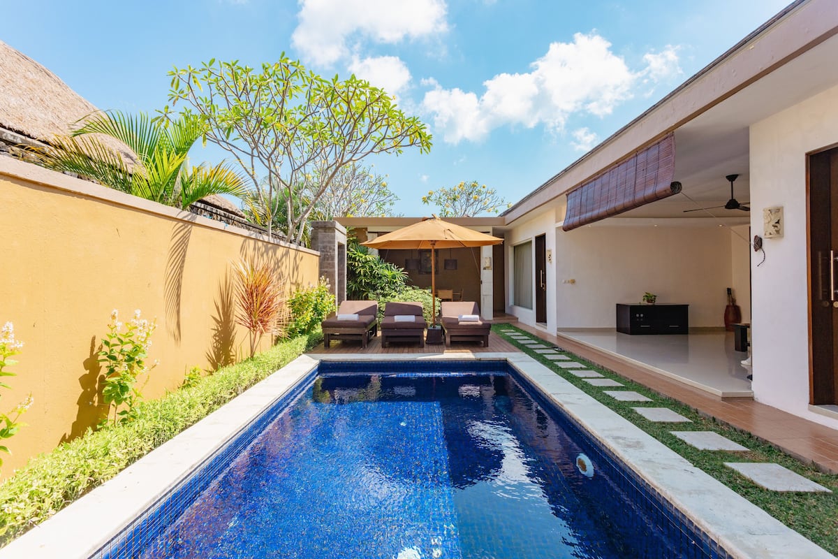 A swimming pool with a sun lounger and a small table, surrounded by a garden and a building with a door and a ceiling fan.