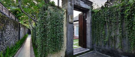 Un vialetto stretto che conduce a un cancello tradizionale, con un muro in pietra da un lato e un fitto siepe verde dall'altro.