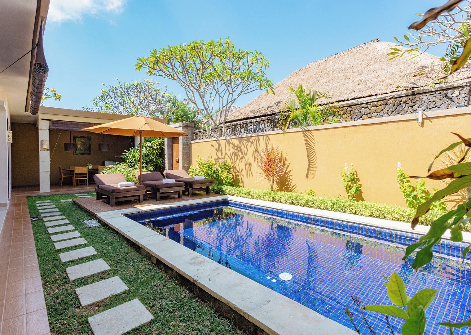 Uma piscina com borda azulejada, rodeada por um pequeno jardim com pedras, um terraço com espreguiçadeiras e uma construção com telhado de palha.