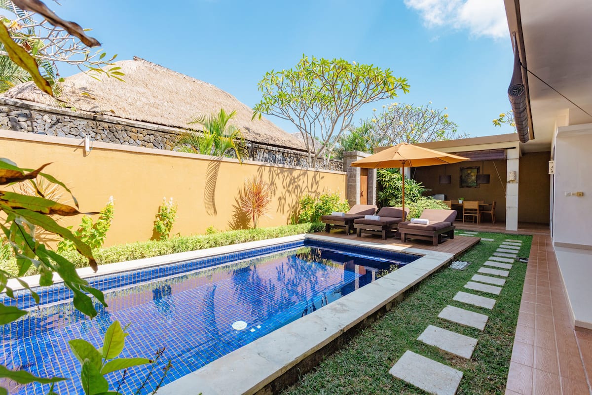 A swimming pool with a tiled edge, a thatched roof structure, and a patio area with lounge chairs and a table.