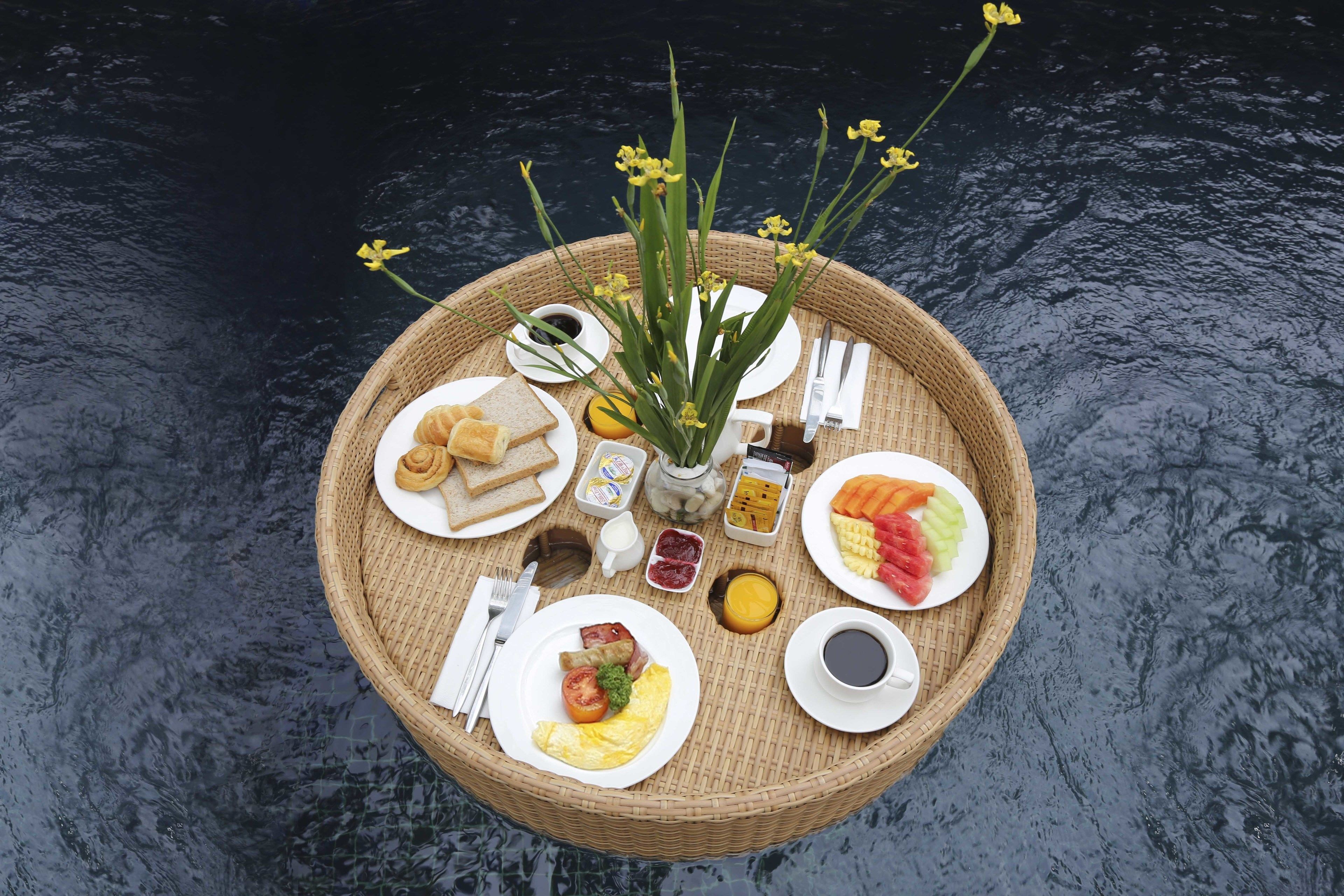 A floating dining setup with various food items and drinks on a round wicker table, surrounded by water.
