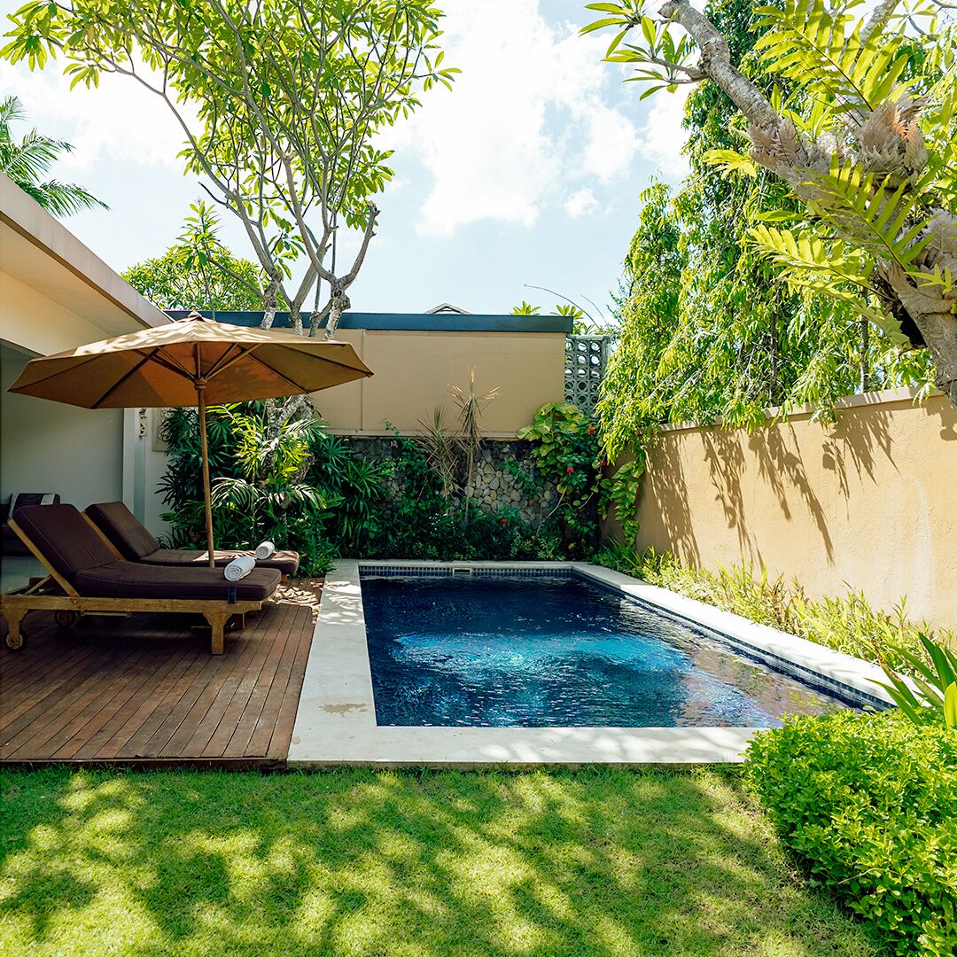 A swimming pool with a wooden deck and lounge chairs, surrounded by lush greenery and a stone wall.