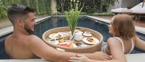 A couple relaxing by a poolside with a wicker tray of food and drinks.