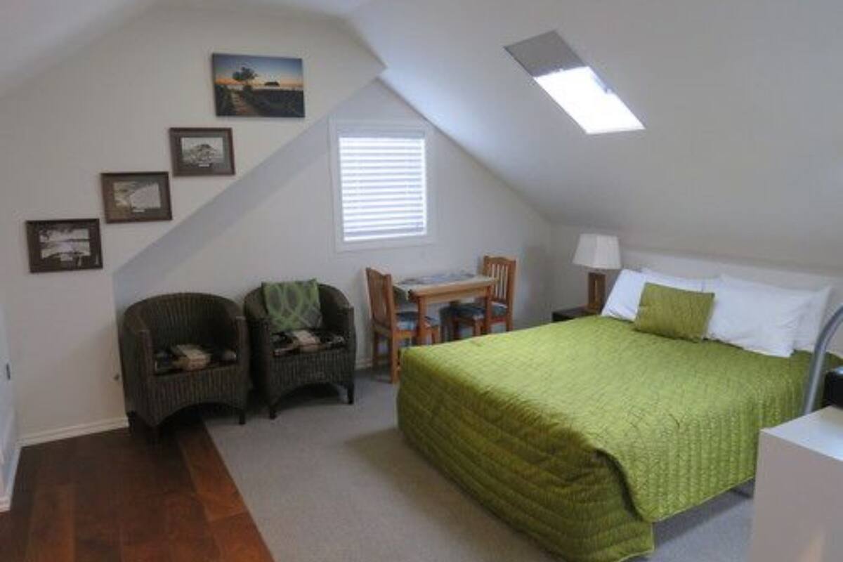 A small, cozy bedroom with a sloped ceiling, a bed with green bedding, a desk with a chair, and a window with blinds.