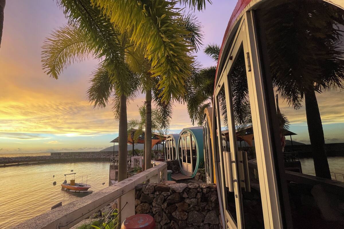 A waterfront view with a stone wall, palm trees, and a boat docked by the water.
