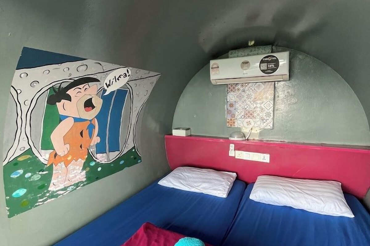 A small, single-bed room with a red and blue bedspread, a white pillow, a blue blanket, and a cartoon drawing on the wall.