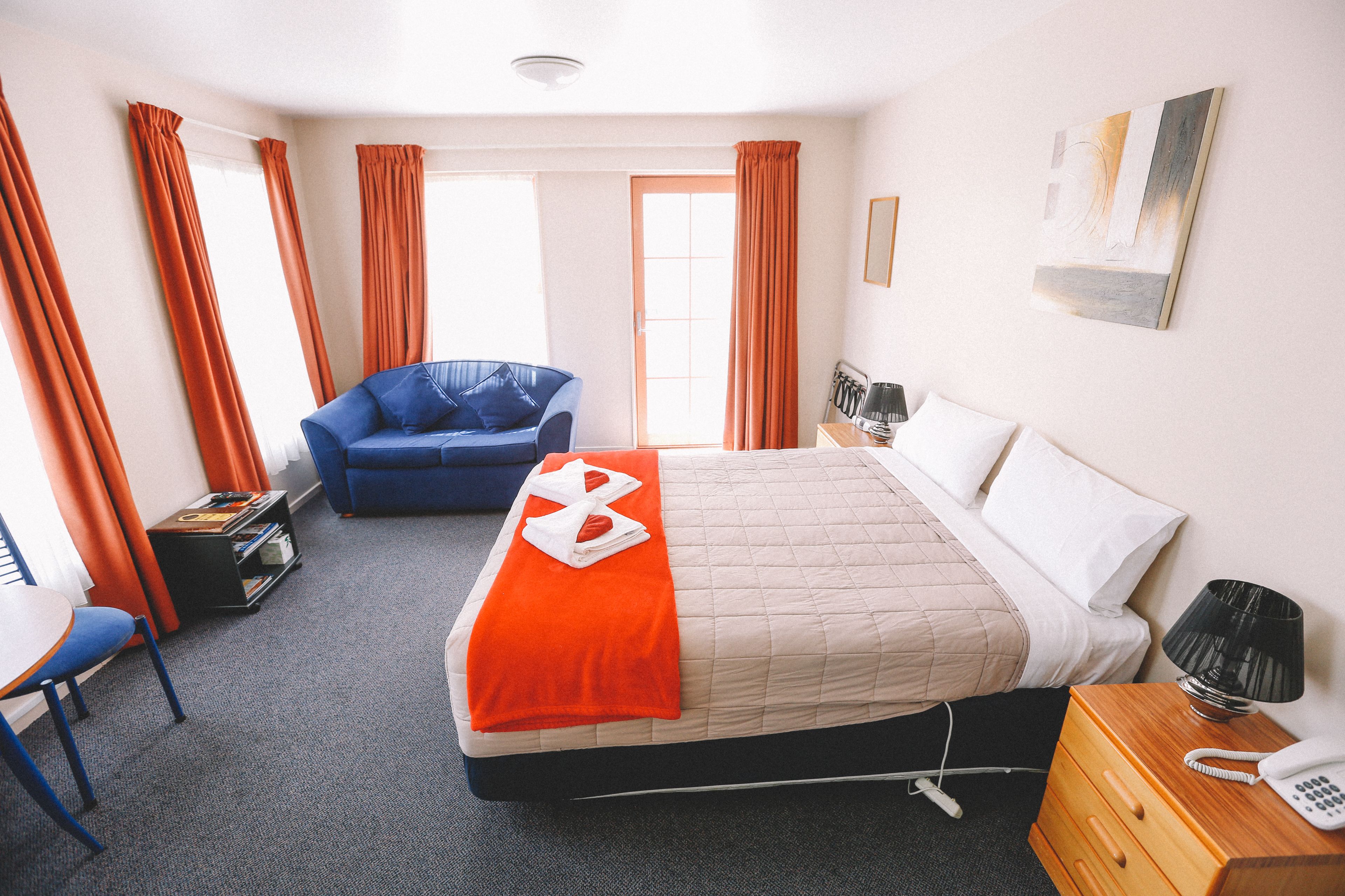 Habitación de hotel con una cama, un sillón azul, un escritorio y una mesita de noche con lámpara y teléfono.
