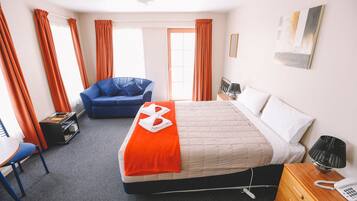 Habitación de hotel con una cama, un sillón azul, un escritorio y una mesita de noche con lámpara y teléfono.