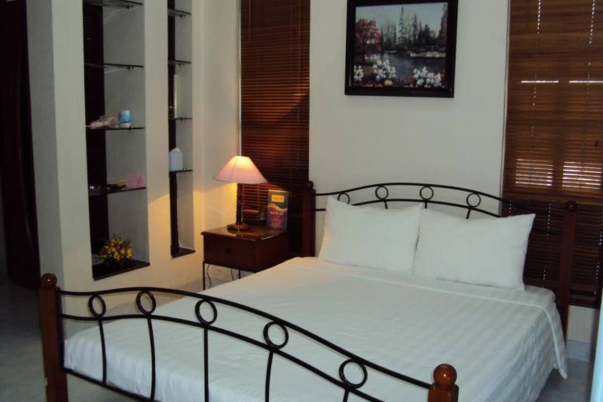 Una cama bien hecha con estructura metálica y ropa de cama blanca, una mesita de noche con una lámpara, un estante con libros y un jarrón con flores, y un cuadro en la pared.