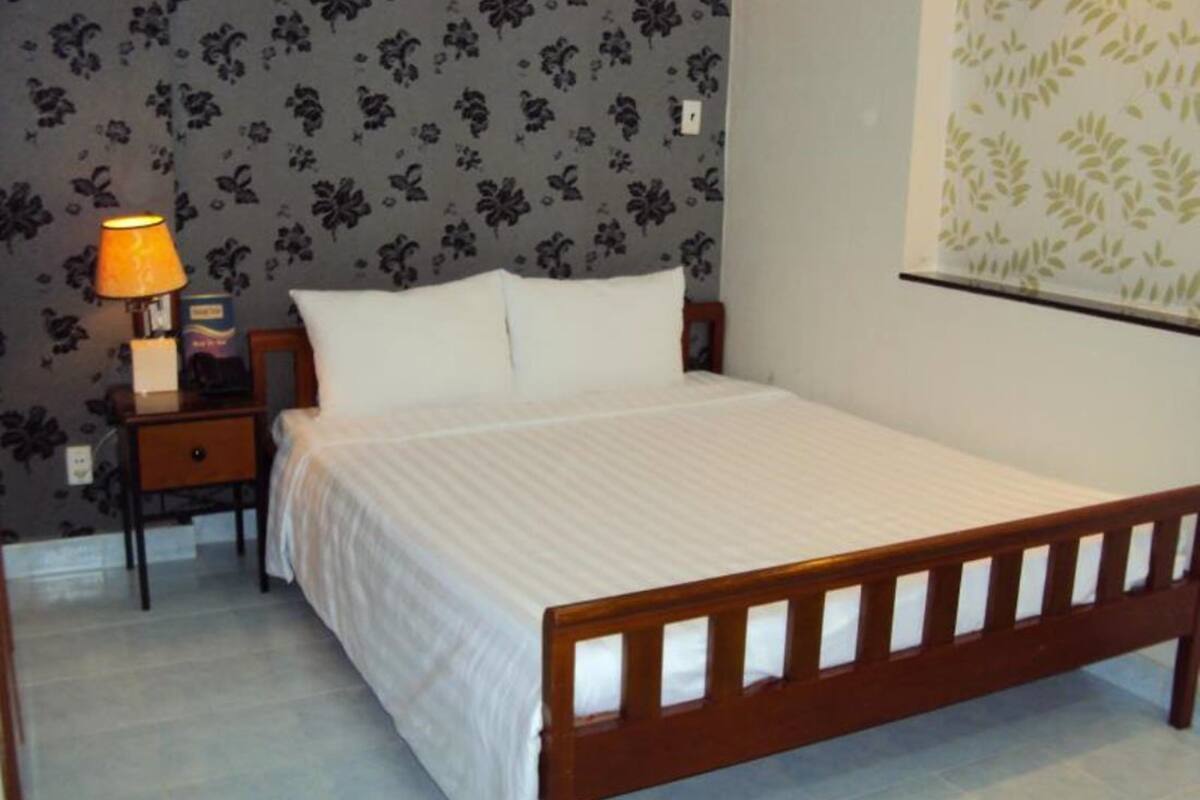 Un dormitorio con cama de madera, ropa de cama blanca, una mesita de noche con lámpara y papel tapiz floral.