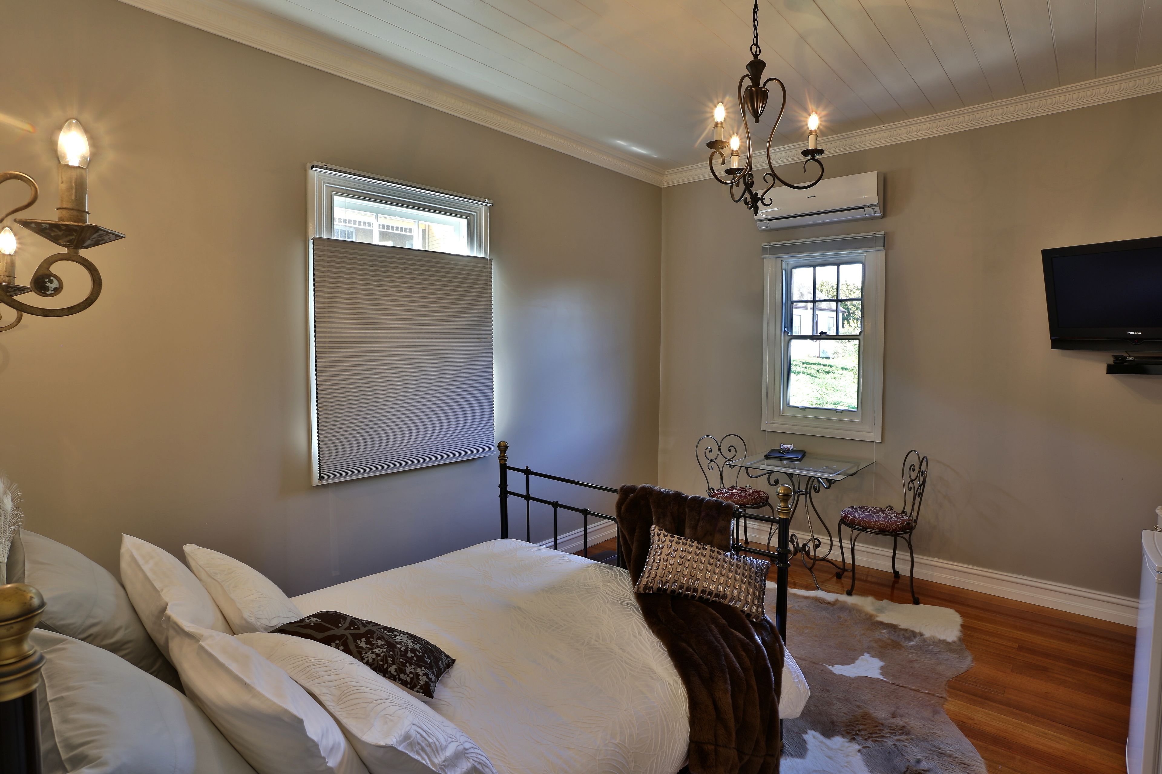 A bedroom with a bed, a chandelier, a television, and a window with blinds.