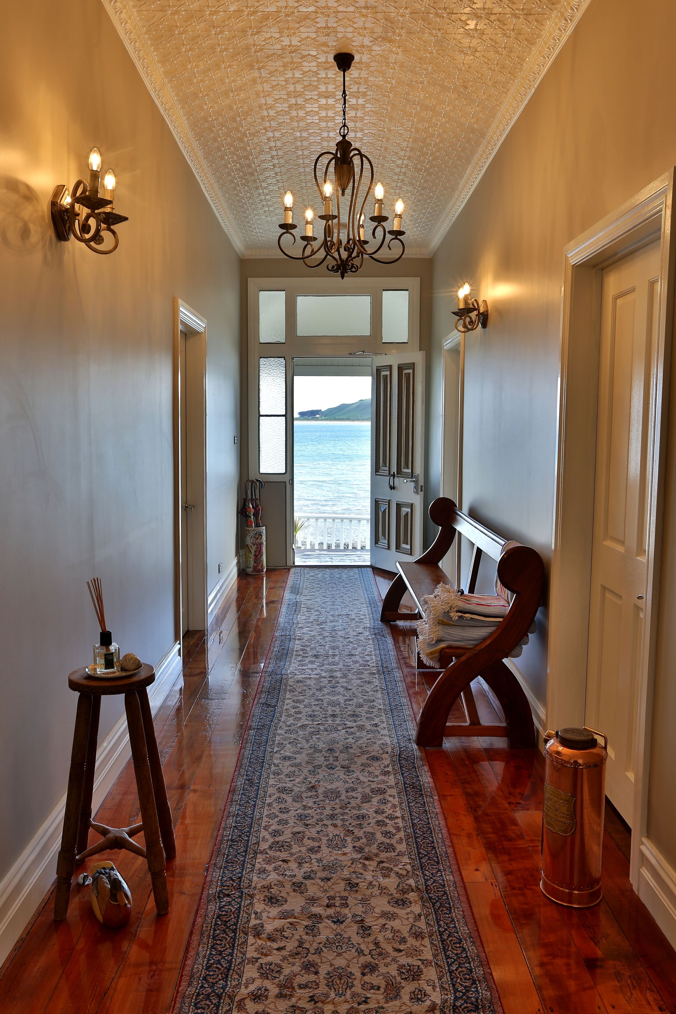 A hallway with a wooden floor, a patterned rug, a bench, a wall-mounted light fixture, and a view of the sea through a glass door.